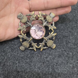 May include: A vintage-style brooch with a central pink gemstone, surrounded by a gold-toned thistle design. The brooch has a square shape and intricate detailing, showcasing a classic and elegant aesthetic. The brooch is a beautiful accessory.