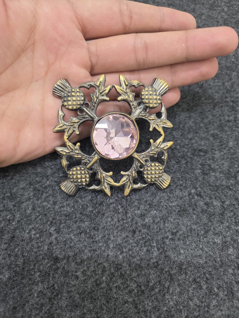 May include: A vintage-style brooch with a central pink gemstone, surrounded by a gold-toned thistle design. The brooch has a square shape and intricate detailing, showcasing a classic and elegant aesthetic. The brooch is a beautiful accessory.