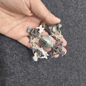 May include: A silver-toned brooch with an ornate, openwork design. The brooch features a central, circular element surrounded by a square frame with stylized floral or leaf-like shapes. A pin and clasp are visible on the back.