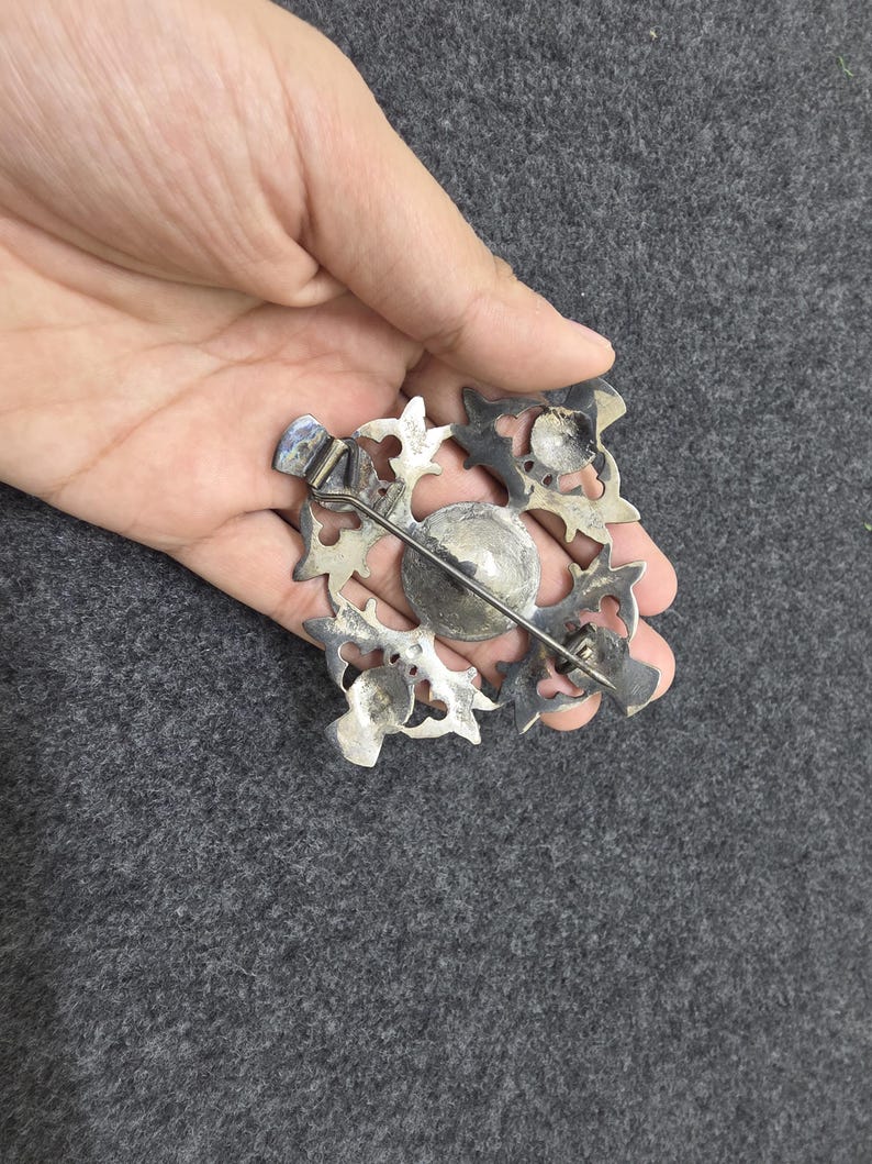 May include: A silver-toned brooch with an ornate, openwork design. The brooch features a central, circular element surrounded by a square frame with stylized floral or leaf-like shapes. A pin and clasp are visible on the back.