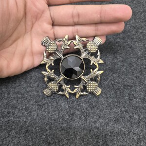 May include: A square brooch with a central black gem, surrounded by a gold-toned thistle and leaf design. The brooch has a vintage aesthetic and is set against a gray background.