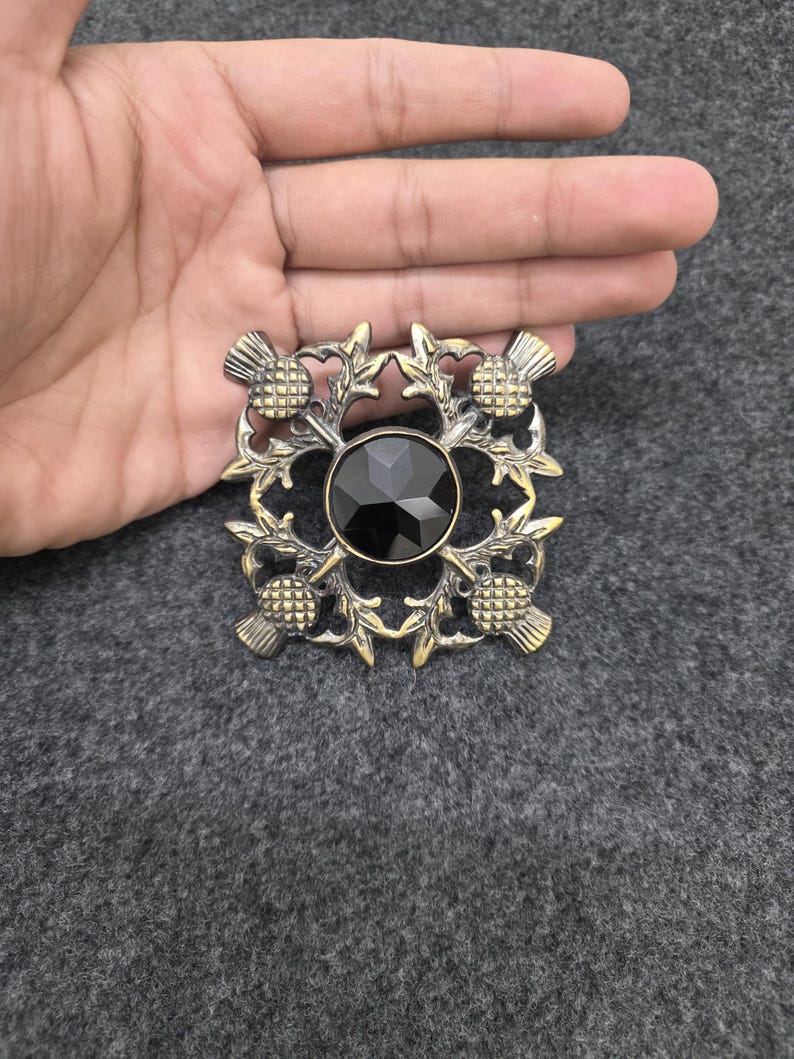 May include: A square brooch with a central black gem, surrounded by a gold-toned thistle and leaf design. The brooch has a vintage aesthetic and is set against a gray background.