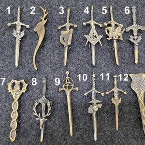May include: A selection of twelve antique brooches, each with a distinct design. The brooches showcase sword, stag, harp, and Masonic emblem motifs, in silver and gold tones. The brooches are displayed on a grey fabric surface.