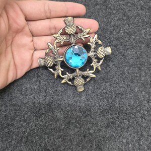 May include: A bronze-toned brooch with a central, faceted, light blue gemstone. The brooch features a thistle design with intricate detailing. The design is circular with four thistles and leaves surrounding the gemstone.