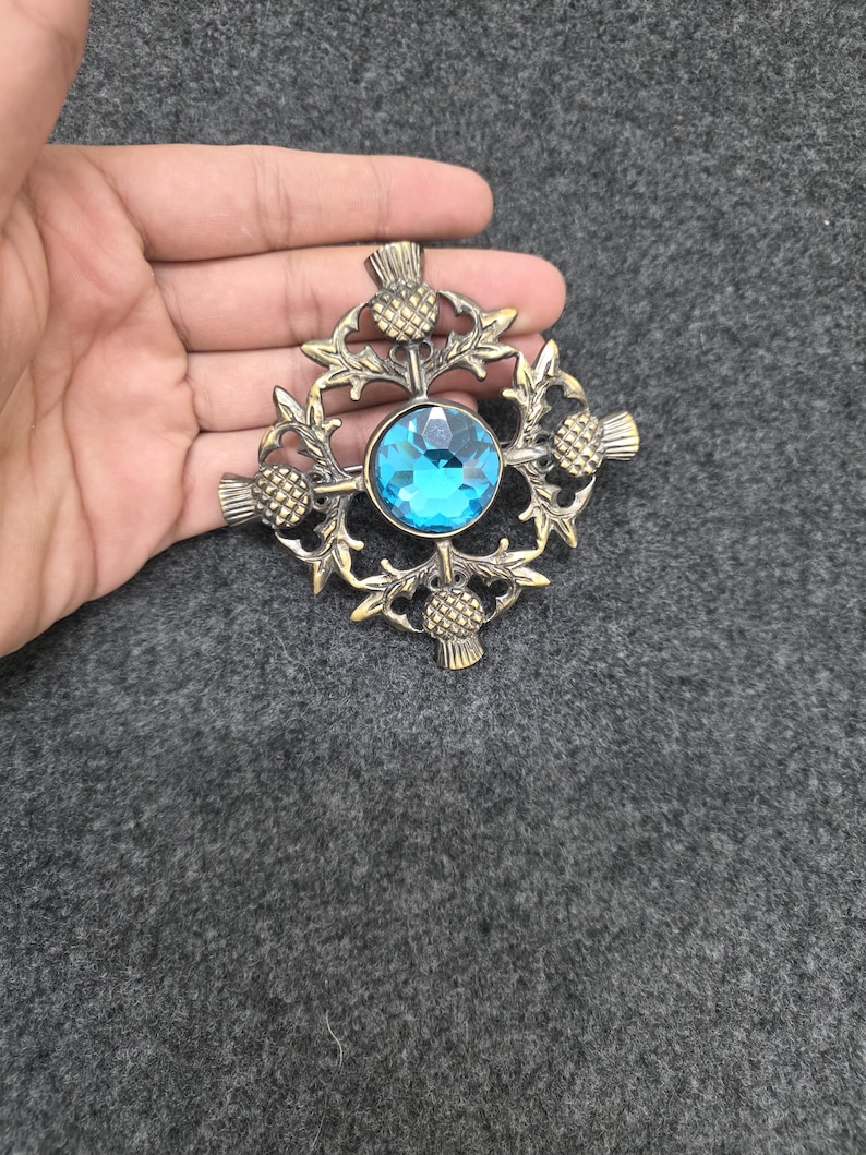 May include: A bronze-toned brooch with a central, faceted, light blue gemstone. The brooch features a thistle design with intricate detailing. The design is circular with four thistles and leaves surrounding the gemstone.