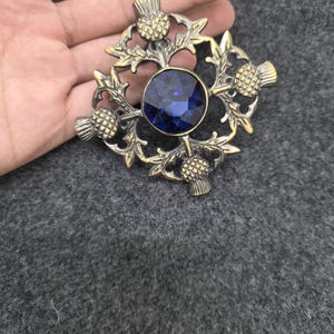 May include: A vintage-style brooch with a central, round, deep blue gemstone. The brooch features an ornate, gold-toned metal design with thistle motifs. The design is symmetrical and detailed, with the gemstone set in the center.