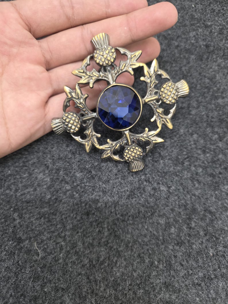May include: A vintage-style brooch with a central, round, deep blue gemstone. The brooch features an ornate, gold-toned metal design with thistle motifs. The design is symmetrical and detailed, with the gemstone set in the center.