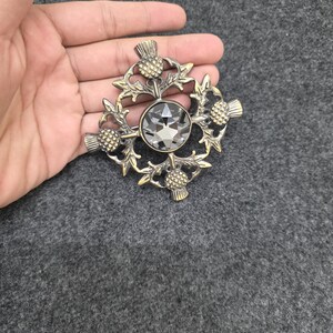 May include: A vintage-style brooch with a central, faceted gray crystal. The brooch features a detailed design of thistles and leaves in a gold-toned metal. The brooch is held in a hand.