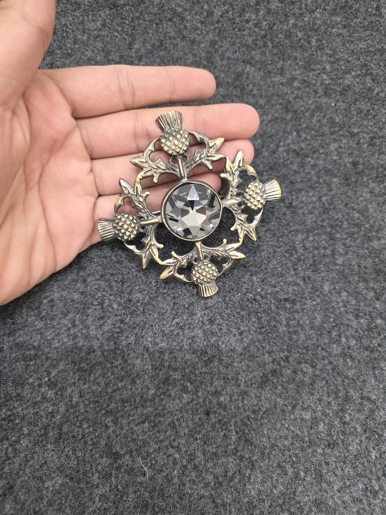 May include: A vintage-style brooch with a central, faceted gray crystal. The brooch features a detailed design of thistles and leaves in a gold-toned metal. The brooch is held in a hand.