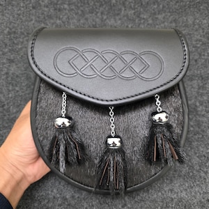 Può includere: Sporran in pelle nera con un motivo a nodo celtico in rilievo sul lembo. Lo sporran presenta un corpo in pelliccia e tre nappe decorative con dettagli in metallo argentato. Un accessorio scozzese tradizionale.