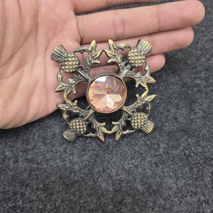 May include: A vintage-style brooch with a central peach-colored gemstone, surrounded by an ornate bronze-toned thistle design. The brooch is square-shaped and features detailed floral and thistle elements, creating a classic, elegant accessory.