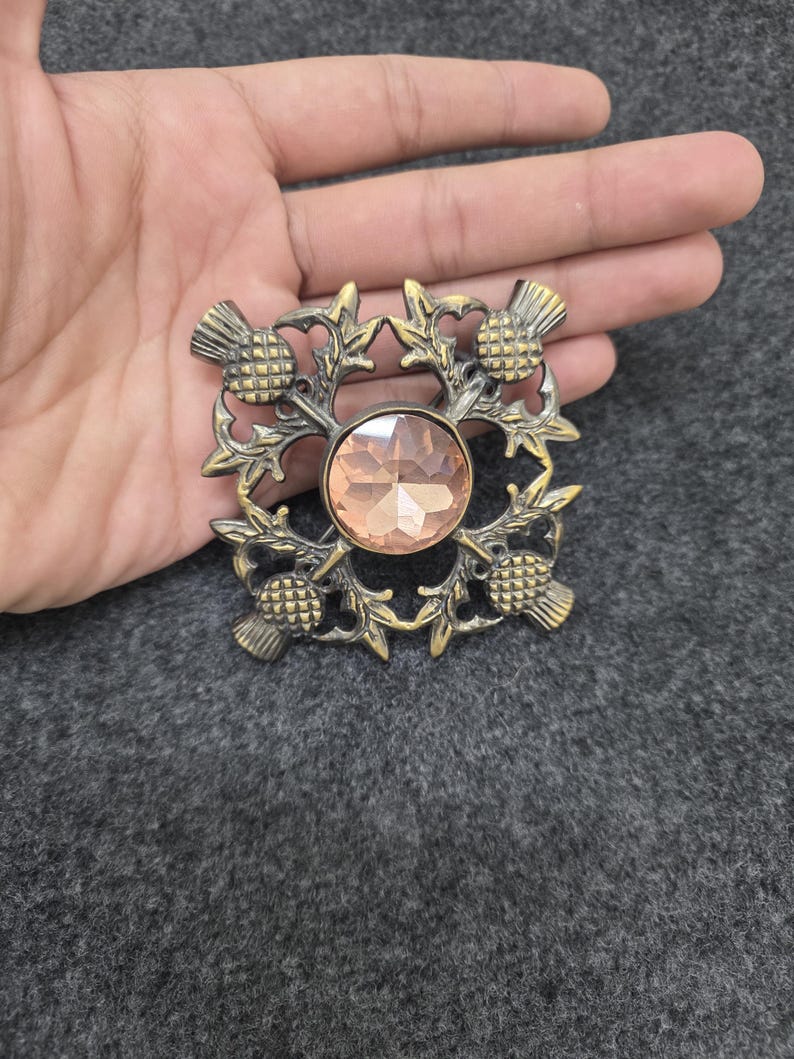 May include: A vintage-style brooch with a central peach-colored gemstone, surrounded by an ornate bronze-toned thistle design. The brooch is square-shaped and features detailed floral and thistle elements, creating a classic, elegant accessory.