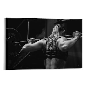 May include: Monochrome image of a person doing a barbell squat in a gym. The person is wearing a sports bra and has their hair in a ponytail. The barbell has weight plates attached, with the word "SUPRFIT" visible.