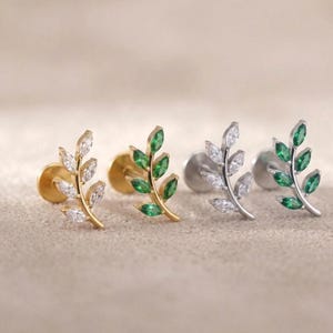 Op de afbeelding: Vier paar blad-vormige oorstekers. Twee paar zijn goudkleurig, één met heldere stenen en de andere met groene stenen. De andere twee paar zijn zilverkleurig, één met heldere stenen en de andere met groene stenen.