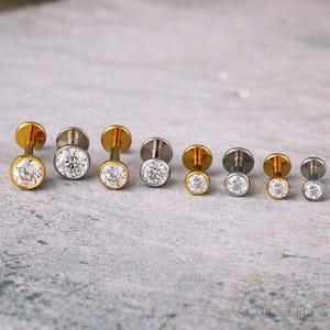 May include: A selection of labret stud earrings with clear, round cubic zirconia stones. The earrings have a flat disc backing and are available in gold and silver tones. The jewellery is displayed on a grey surface.