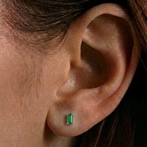 May include: A close-up of an ear with a small, rectangular emerald stud earring. The emerald is a vibrant green, set in a gold-colored metal. The earring is positioned on the earlobe.
