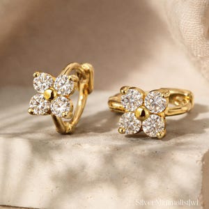 May include: A pair of gold-coloured hoop earrings. Each earring features a four-petal flower design, with clear, sparkling stones set in each petal. A small, gold-coloured bead is at the centre of each flower. The earrings are displayed on a neutral surface.