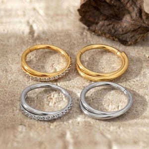 Può includere: Quattro orecchini circolari. Due sono dorati, uno con una fila di piccole pietre trasparenti, l'altro con un design liscio a doppia fascia. Gli altri due sono argentati, uno con pietre e l'altro con un design a doppia fascia.