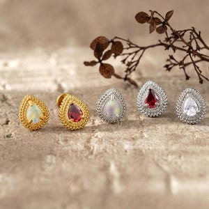 Può includere: Un assortimento di orecchini a lobo a forma di goccia. Due sono dorati con un bordo strutturato, uno con un'opale bianca e l'altro con una gemma rossa. Gli altri tre sono argentati con un bordo strutturato, ognuno con una gemma diversa.
