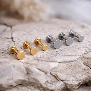 Peut inclure: Une sélection de boucles d'oreilles à clous à dos plat, de tons or et argent. Les boucles d'oreilles ont une face circulaire et une tige droite. Les boucles d'oreilles sont présentées sur une surface texturée de couleur claire.