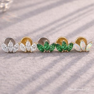 Puede incluir: Cuatro pendientes de botón con diseño de hoja, con gemas de corte marquesa. Los pendientes son de oro y plata, con gemas blancas, verdes y ópalo. Se muestran sobre una superficie gris.