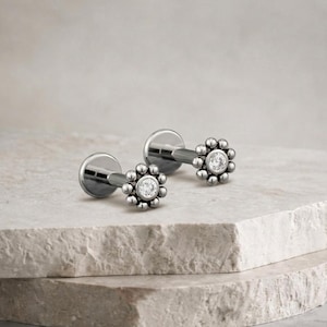 Puede incluir: Dos pendientes plateados con forma de flor, cada uno con una gema transparente central y una corona de pequeñas cuentas. Los pendientes se exhiben sobre una superficie clara y texturizada, resaltando su diseño delicado y brillo.