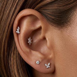 Op de afbeelding: Close-up van een oor versierd met meerdere sieraden. Het oor heeft drie kleine, sprankelende oorbellen. Eén is een kleine, ronde knop, een andere heeft een vlindervorm en de derde is een bloemenontwerp.