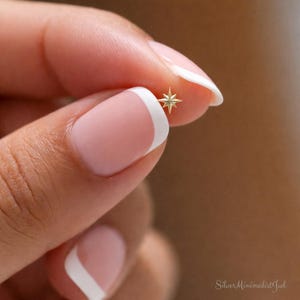 Puede incluir: Un pequeño pendiente dorado en forma de estrella sostenido entre los dedos. El pendiente tiene ocho puntas y un diseño minimalista. La imagen es un primer plano, que destaca los delicados detalles del pendiente y el tono de piel suave de la mano.