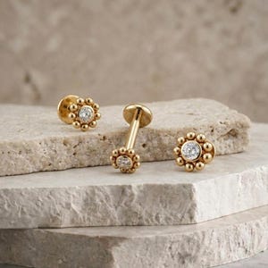 Puede incluir: Tres pendientes dorados con forma de flor y una gema transparente en el centro. Los pendientes se muestran sobre una superficie clara y texturizada. El diseño presenta pequeños detalles redondos alrededor de la piedra central.