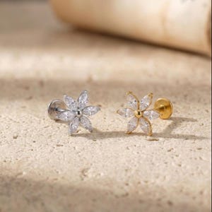 Puede incluir: Dos pendientes de botón con forma de flor. Uno es plateado y el otro dorado. Cada pendiente presenta una gema central redonda rodeada de seis gemas en forma de marquesa, creando un diseño floral. Los pendientes se muestran sobre una superficie clara y texturizada.