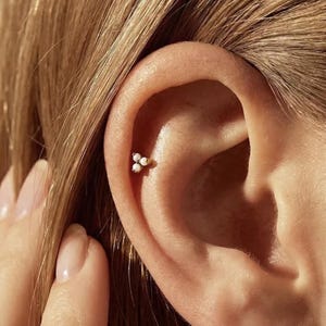 May include: A close-up of an ear with a small, gold-coloured earring. The earring features three small, white, pearl-like beads arranged in a triangular shape. The skin around the ear is light-toned, and the hair is a light brown colour.