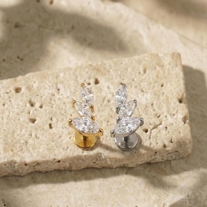 Puede incluir: Dos pendientes de botón, uno dorado y otro plateado, cada uno con un grupo de piedras preciosas claras con corte marquesa. Las piedras están dispuestas en forma de hoja. Los pendientes se muestran sobre una superficie texturizada de color marrón claro.