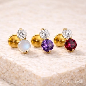 Peut inclure: Trois boucles d'oreilles à clous dorées, chacune avec une petite gemme blanche scintillante au-dessus d'une pierre précieuse colorée plus grande. Les pierres précieuses sont bleu clair, violet et rouge. Les boucles d'oreilles sont présentées sur une surface claire.