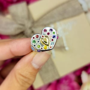 May include: A heart-shaped ring with a silver band and a gold heart in the center. The ring is encrusted with colorful gemstones. The ring is held in a hand, with a blurred background of pink flowers and a gift box.