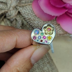 May include: Silver heart-shaped ring with a flower and gemstone design. The ring features a gold flower with a red center and is surrounded by various colorful gemstones. The ring is held in a hand, with a blurred background of a gift box and pink flower.