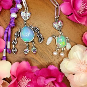 May include: Two whimsical, handmade pendant necklaces with iridescent oval stones and silver-toned metal accents. Each necklace features a heart-shaped face charm and is adorned with dangling charms and pearls. The background is decorated with pink and white flowers.
