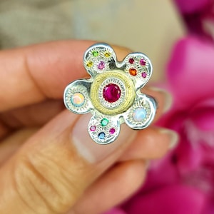 May include: A silver flower-shaped ring featuring a central round red gemstone set in a gold-toned textured circle. The petals are accented with small, multi-colored gemstones including opals, rubies, and sapphires, creating a vibrant, detailed design.