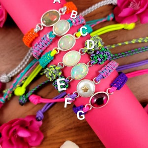Puede incluir: Primer plano de siete pulseras con cordones de diferentes colores y dijes de plata. Cada dije presenta una gema diferente, incluyendo ópalo y un dije en forma de estrella. Las pulseras están dispuestas en una pila sobre una superficie rosa.