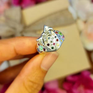 May include: A silver ring with a bow design, adorned with a variety of colorful gemstones. The ring features a detailed, textured surface and is set against a blurred background of pink flowers and a gift box.