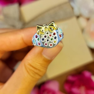 May include: A silver ring with a light blue band and a gold bow on top. The ring is adorned with colorful gemstones, including red, green, and blue stones. The ring is held in a person's hand.