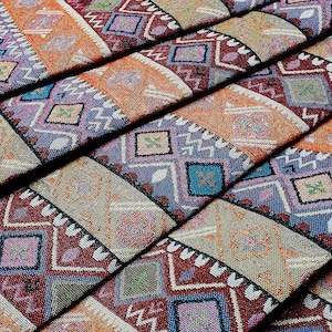 May include: Close-up of a stack of fabric with a colorful geometric pattern. The fabric is woven with a variety of colors, including red, orange, blue, and green. The pattern is made up of repeating diamonds and triangles.