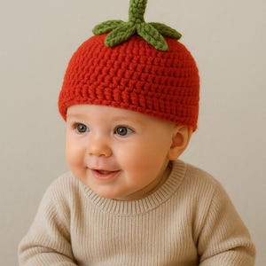 Puede incluir: Un bebé con un gorro de tomate de ganchillo rojo hecho a mano con hojas verdes. El bebé también lleva un suéter beige. El gorro es un accesorio divertido y único para un bebé.
