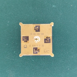 Könnte beinhalten: Ein quadratisches, goldfarbenes elektronisches Bauteil mit vier kleinen schwarzen rechteckigen Kontaktpunkten, die um ein zentrales Loch angeordnet sind. Die Oberfläche ist mit 76 426 046 060, D 824 T Z 32 und MC beschriftet.