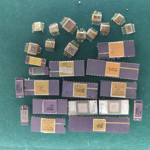 May include: A collection of various electronic integrated circuit chips in different sizes, featuring dark purple and metallic gold rectangular bodies with multiple metal pins extending from the sides, displaying printed text and serial numbers.