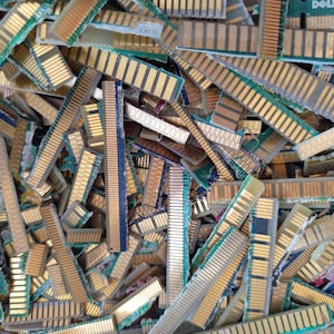 Puede incluir: Una gran colección de módulos de memoria RAM de computadora desechados apilados en un montón caótico. Cada módulo rectangular presenta una base de placa de circuito verde con una fila de pines de contacto de color dorado en un borde.