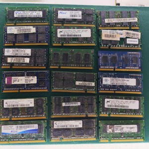 May include: A collection of twenty rectangular computer RAM memory modules arranged in a grid. Each module features a green or blue circuit board with integrated chips, gold-plated contact pins along the bottom edge, and white identification labels.