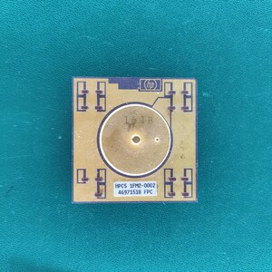 Könnte beinhalten: Eine quadratische, goldfarbene elektronische Komponente mit einem zentralen kreisförmigen Kontakt, dem HP-Logo und einem weißen Etikett mit dem Text HPC5 1FM2-0002 46971518 FPC. Die Oberfläche zeigt dunkelbraune Leiterbahnen und die Zahl 1618.