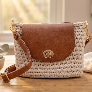Könnte beinhalten: Beige gehäkelte Umhängetasche mit braunem Lederklappe und Riemen. Die Tasche hat goldfarbene Hardware und einen sicheren Verschluss. Die Tasche liegt auf einer Holzoberfläche.
