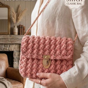 Könnte beinhalten: Eine rosa Häkeltasche mit einem Noppenmuster und einem goldfarbenen Verschluss. Die Tasche hat einen Kettenriemen. Der Text "CROCHET PATTERN PDF DOWNLOAD" und "BOBBLE STITCH CLASSIC BAG" sind sichtbar.