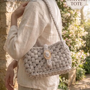 Könnte beinhalten: Eine hellgraue, grob strukturierte Tragetasche mit geflochtenem Riemen und goldfarbenem Verschluss. Die Tasche besteht aus einem dicken, gewebten Material. Das Bild enthält den Text "CROCHET PATTERN PDF DOWNLOAD" und "CHUNKY TEXTURE TOTE". Das Visual Mints-Logo befindet sich unten.